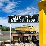 a tall golden, railraod spike on display with two trains in the background